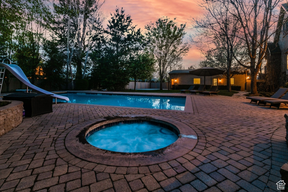 Pool at dusk featuring an in ground hot tub, a water slide, and a patio