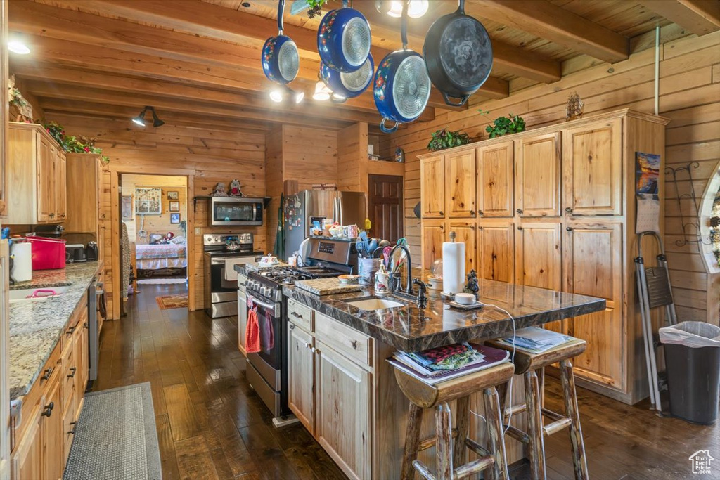 Kitchen featuring light stone counters, dark hardwood / wood-style floors, stainless steel appliances, a center island with sink, and beam ceiling