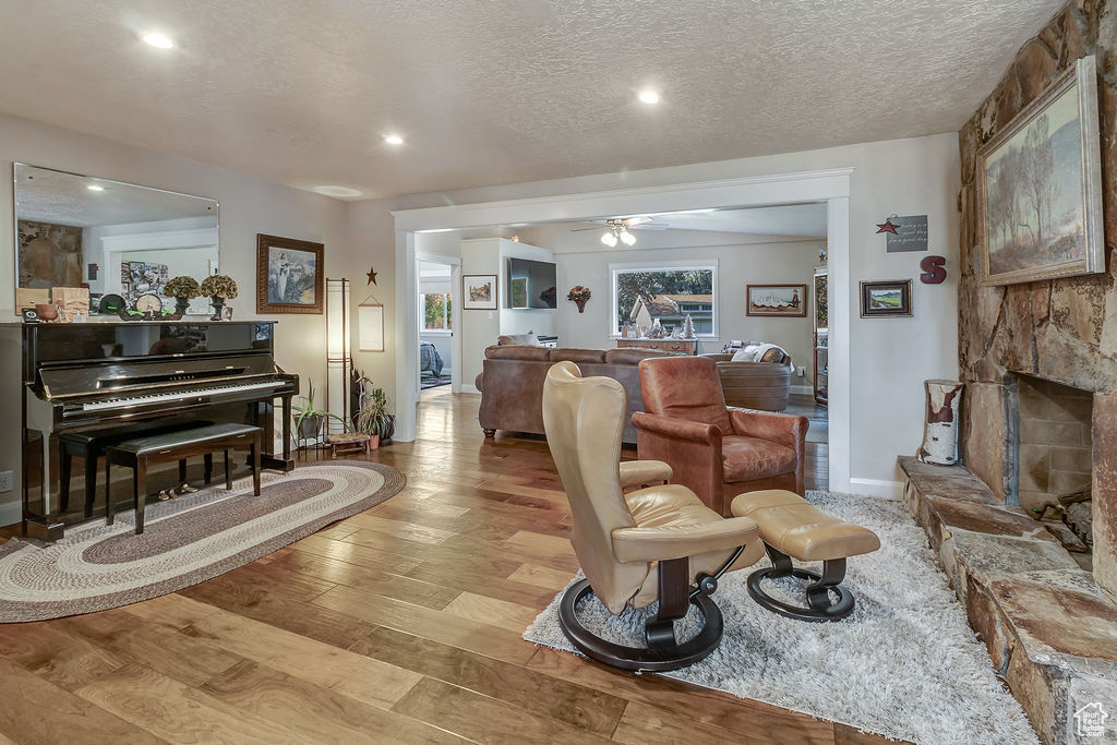 Living room featuring ceiling fan, a stone fireplace, a textured ceiling, and light hardwood / wood-style floors