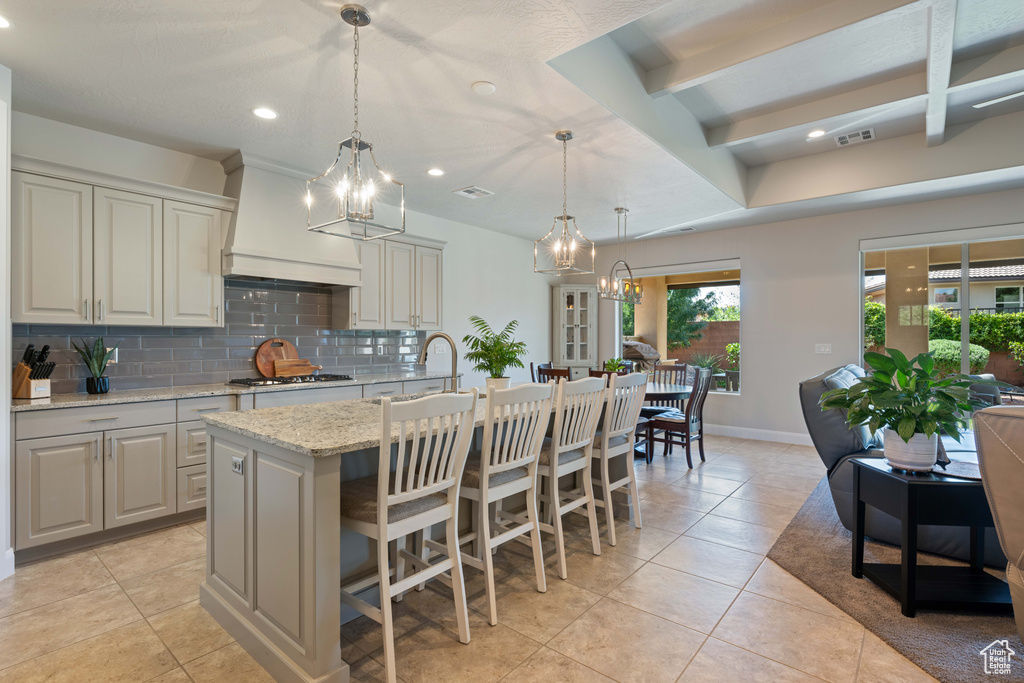 Kitchen featuring pendant lighting, a center island with sink, light stone counters, custom exhaust hood, and backsplash