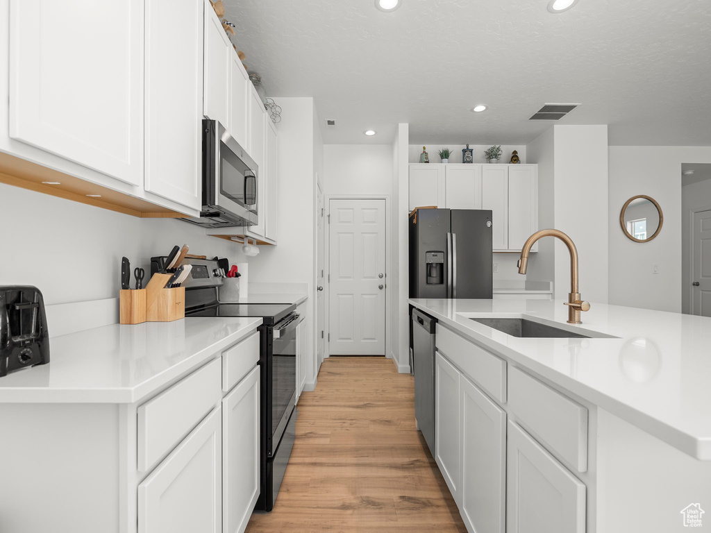 Kitchen with light wood-type flooring, stainless steel appliances, sink, white cabinetry, and a center island with sink