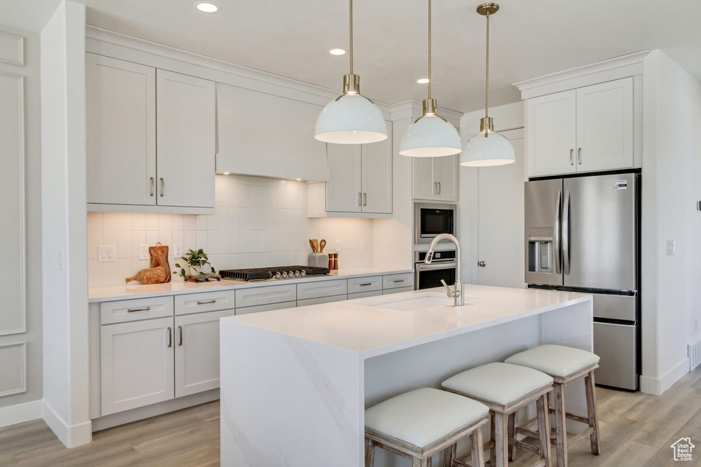 Kitchen with light wood-type flooring, a kitchen island with sink, stainless steel appliances, white cabinetry, and decorative light fixtures