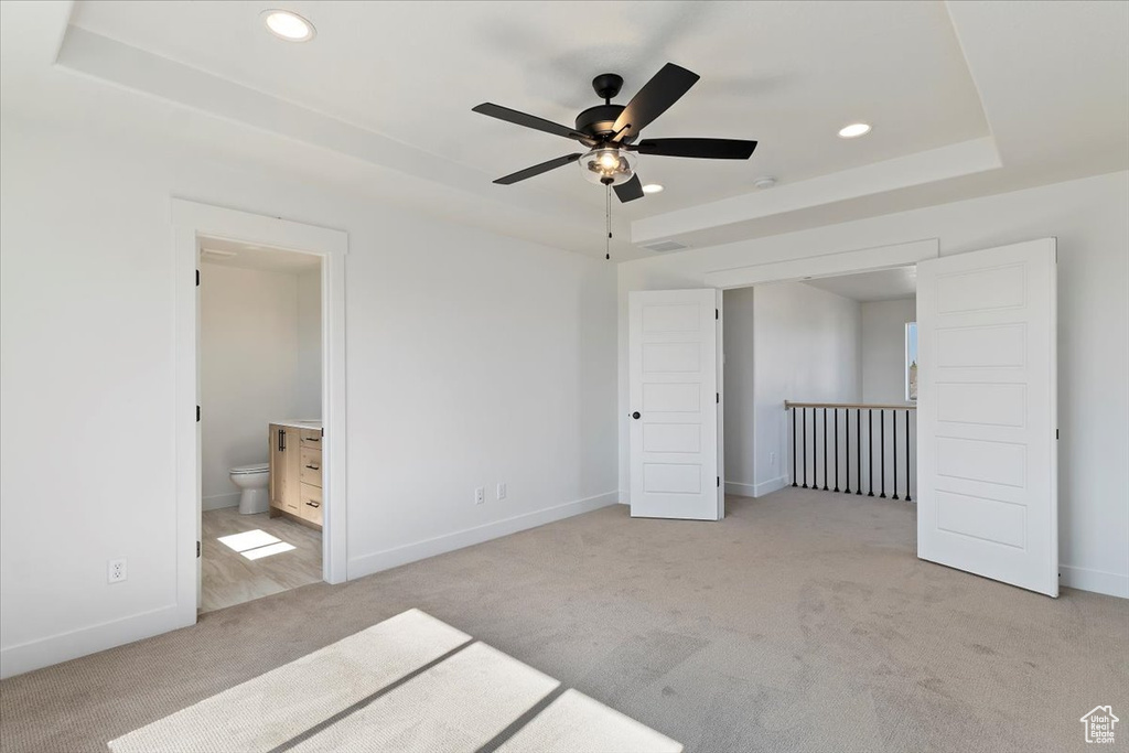 Unfurnished bedroom with a raised ceiling, light colored carpet, connected bathroom, and ceiling fan