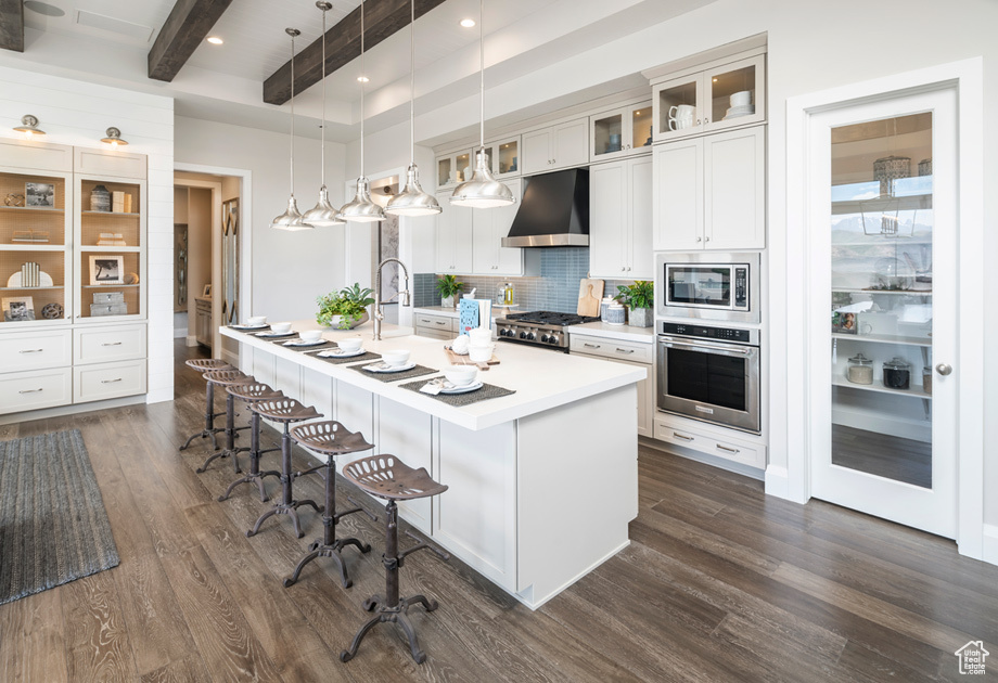 Kitchen with hanging light fixtures, beam ceiling, wall chimney exhaust hood, stainless steel appliances, and dark hardwood / wood-style flooring