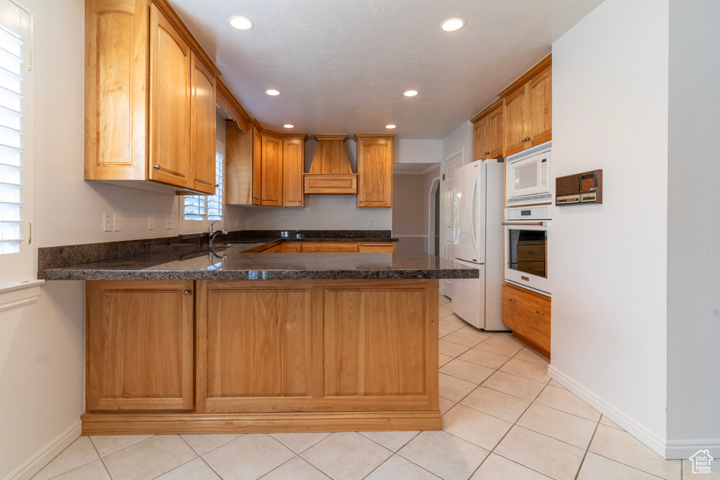 Kitchen featuring premium range hood, white appliances, sink, light tile patterned flooring, and kitchen peninsula