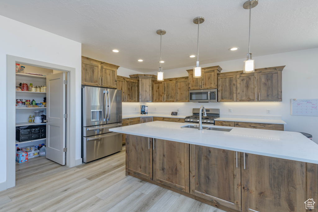 Kitchen featuring appliances with stainless steel finishes, sink, light hardwood / wood-style floors, decorative light fixtures, and a center island with sink