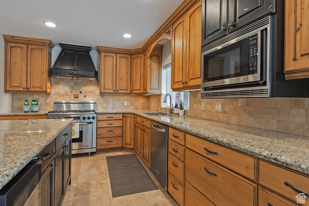 Kitchen with decorative backsplash, light stone counters, appliances with stainless steel finishes, custom range hood, and sink