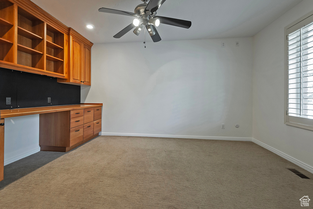 Unfurnished office featuring carpet floors, built in desk, plenty of natural light, and ceiling fan