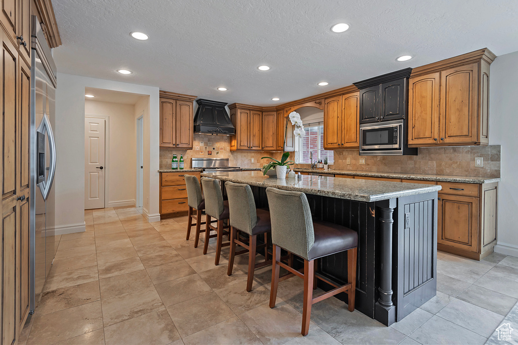 Kitchen featuring light stone countertops, a kitchen island, a kitchen breakfast bar, custom exhaust hood, and built in appliances