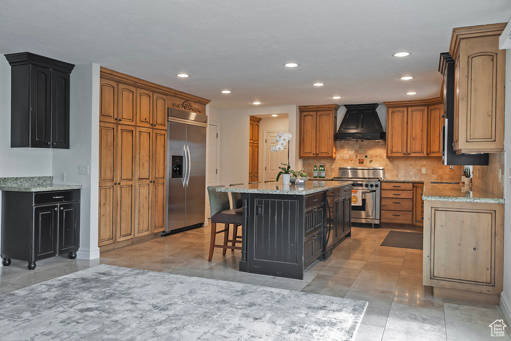 Kitchen with appliances with stainless steel finishes, premium range hood, light stone countertops, and a center island with sink