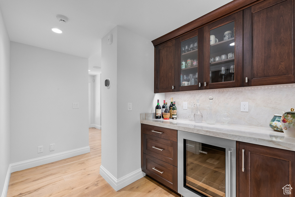 Bar with wine cooler, light stone countertops, dark brown cabinetry, light hardwood / wood-style floors, and tasteful backsplash