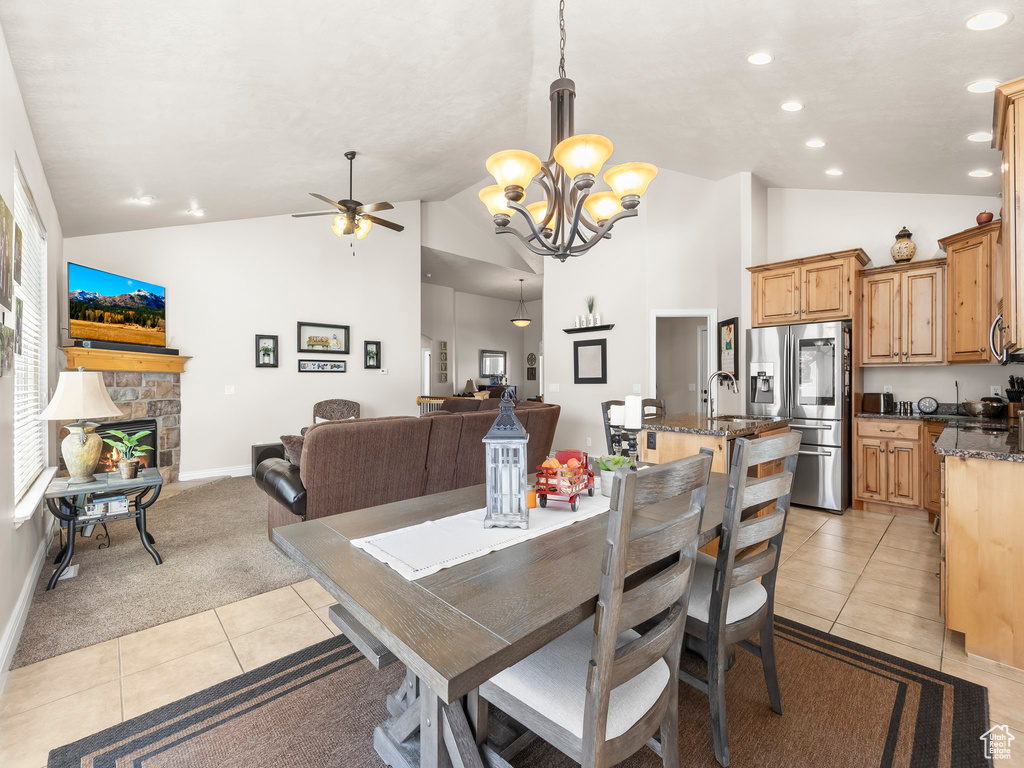 Dining space featuring sink, a stone fireplace, high vaulted ceiling, light carpet, and ceiling fan with notable chandelier