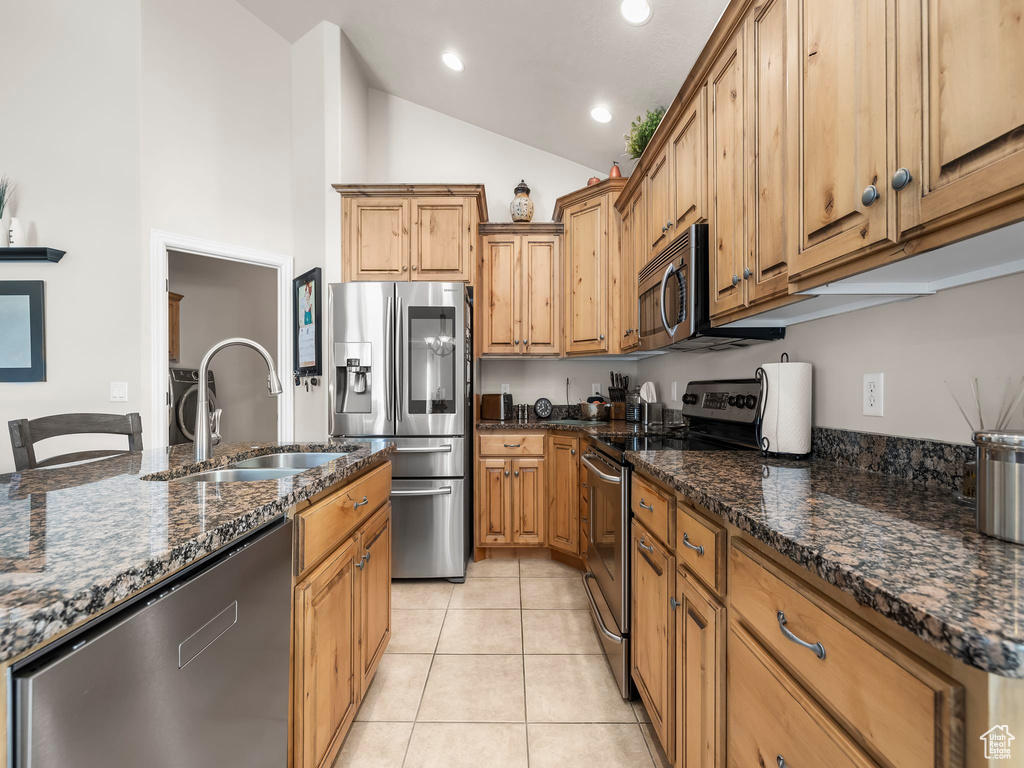 Kitchen featuring light tile patterned flooring, dark stone countertops, vaulted ceiling, sink, and appliances with stainless steel finishes