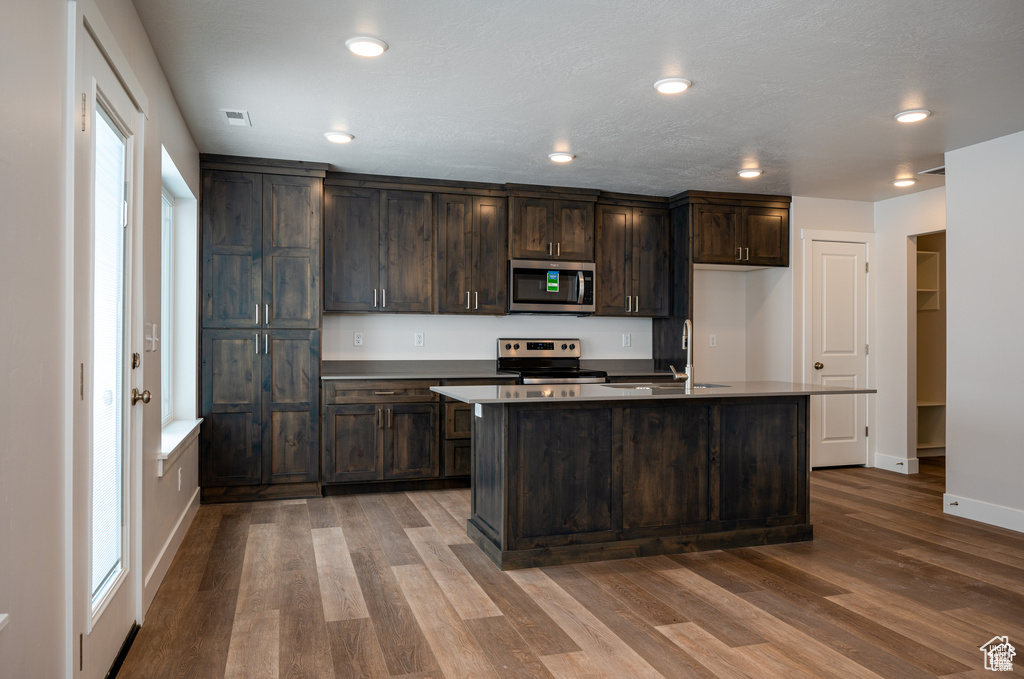 Kitchen with hardwood / wood-style flooring, stainless steel appliances, dark brown cabinets, and a center island with sink