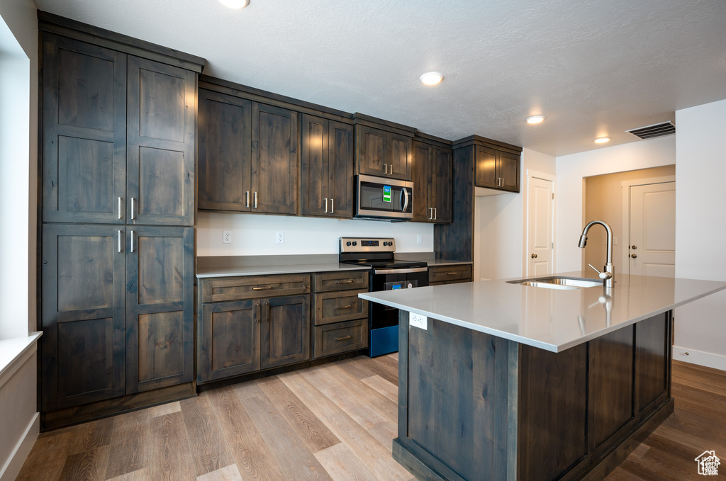 Kitchen featuring dark brown cabinets, a center island with sink, sink, light wood-type flooring, and appliances with stainless steel finishes