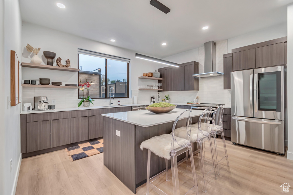 Kitchen with wall chimney range hood, light wood-type flooring, a kitchen island, hanging light fixtures, and stainless steel refrigerator