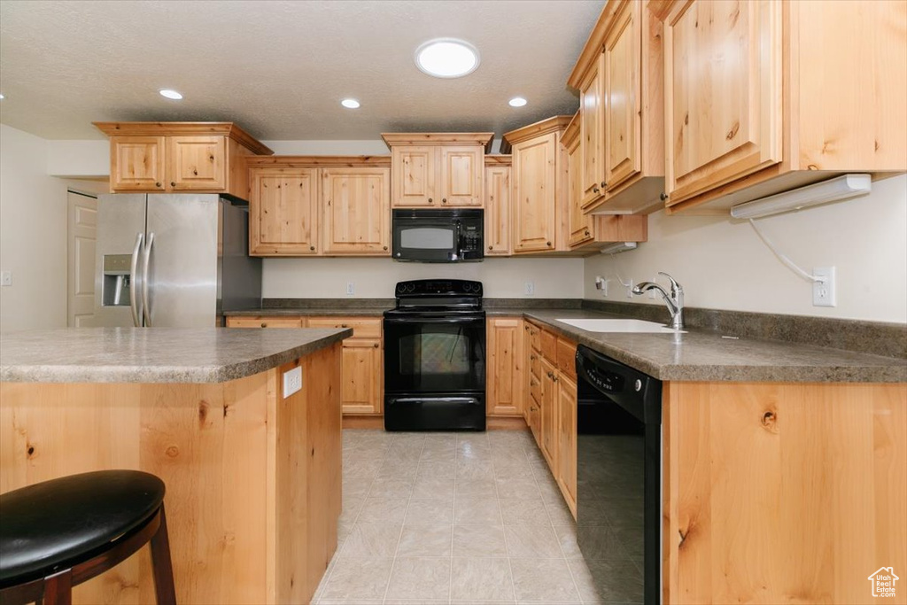 Kitchen with black appliances, a sink, light brown cabinetry, dark countertops, and a breakfast bar