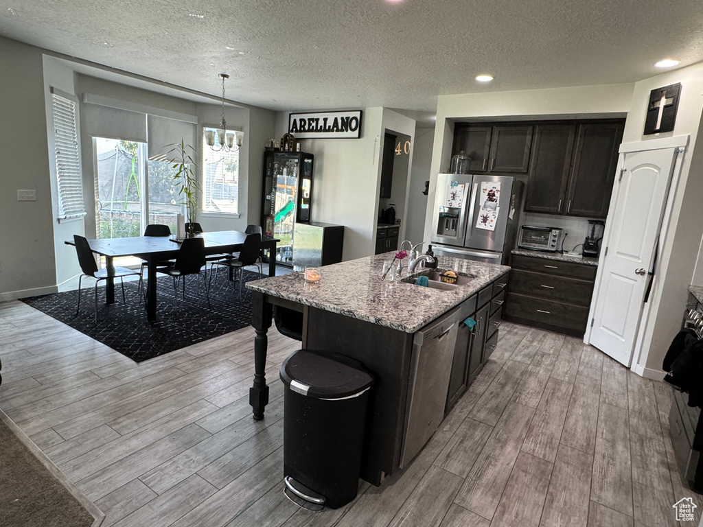 Kitchen with hanging light fixtures, appliances with stainless steel finishes, a kitchen island with sink, light hardwood / wood-style floors, and sink