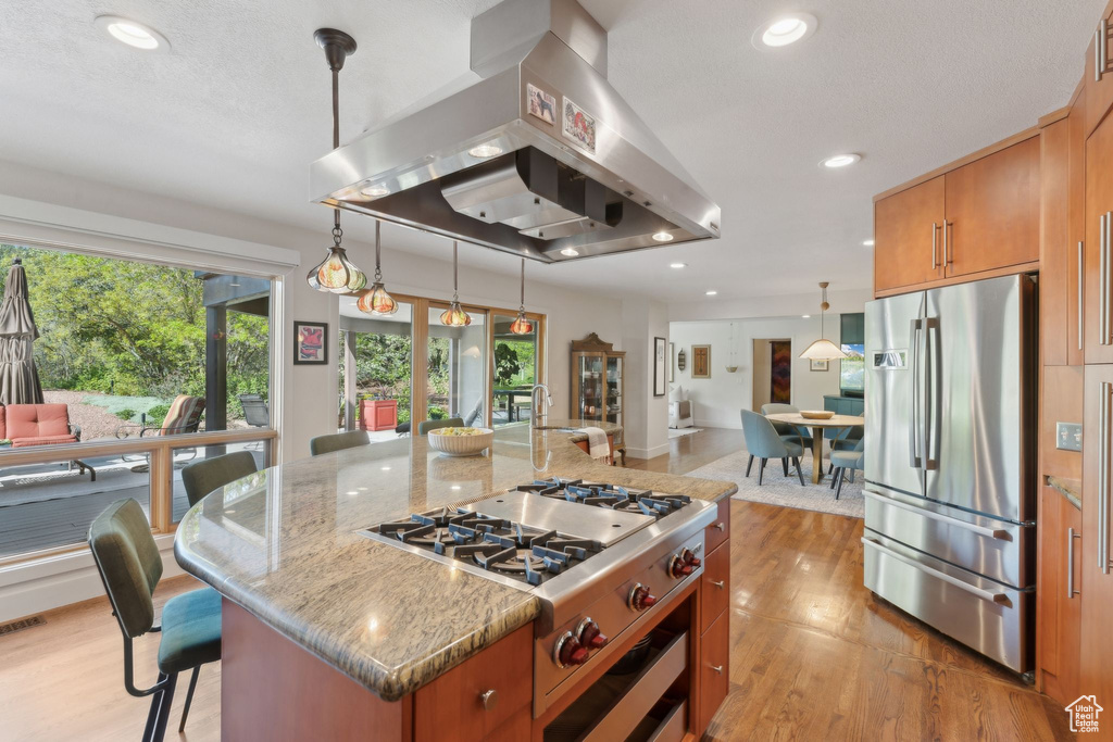 Kitchen featuring appliances with stainless steel finishes, island range hood, light wood-type flooring, brown cabinetry, and light stone counters