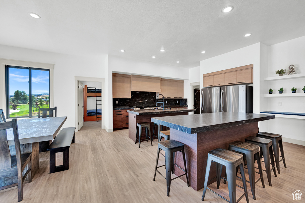 Kitchen with stainless steel refrigerator, tasteful backsplash, light hardwood / wood-style floors, a kitchen island with sink, and a breakfast bar