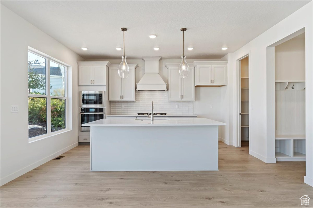 Kitchen with white cabinetry, appliances with stainless steel finishes, custom exhaust hood, and a kitchen island with sink