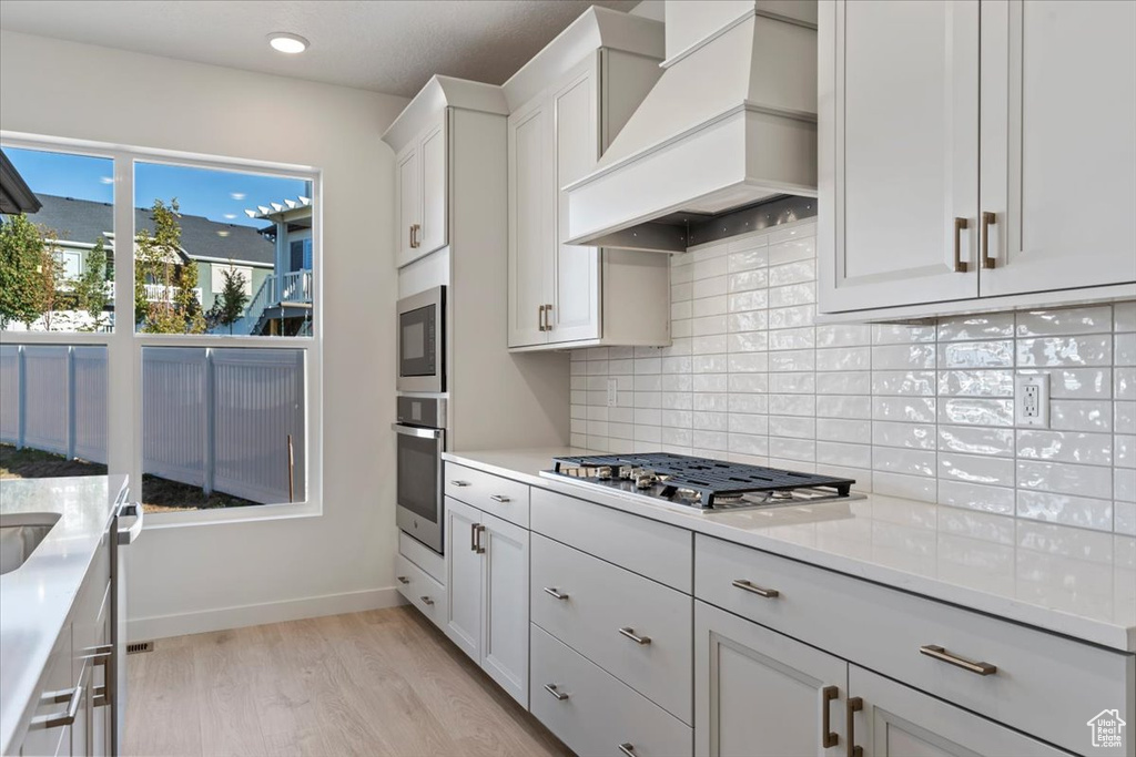 Kitchen featuring custom exhaust hood, white cabinets, tasteful backsplash, light wood-type flooring, and stainless steel appliances