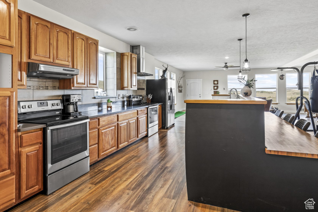 Kitchen featuring appliances with stainless steel finishes, ceiling fan, wall chimney exhaust hood, pendant lighting, and dark wood-type flooring