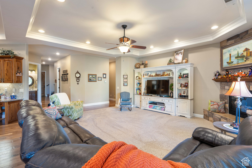 Living room featuring ceiling fan, wood-type flooring, a tray ceiling, ornamental molding, and a stone fireplace