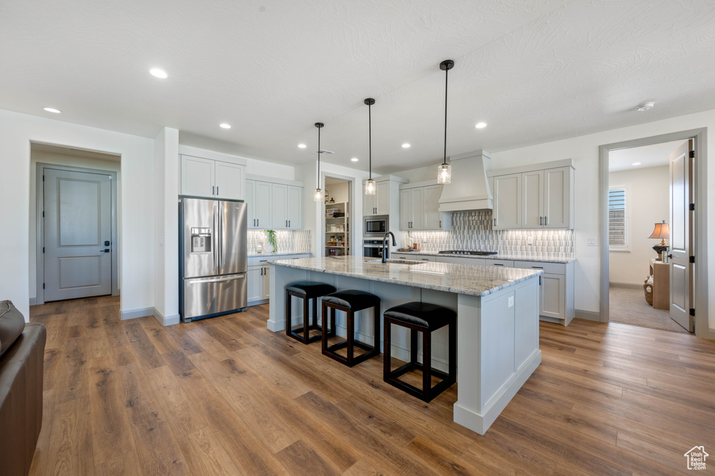 Kitchen with hardwood / wood-style floors, an island with sink, appliances with stainless steel finishes, white cabinetry, and custom range hood