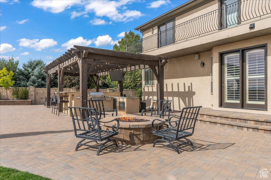 View of patio featuring exterior kitchen, a bar, an outdoor fire pit, a pergola, and a balcony