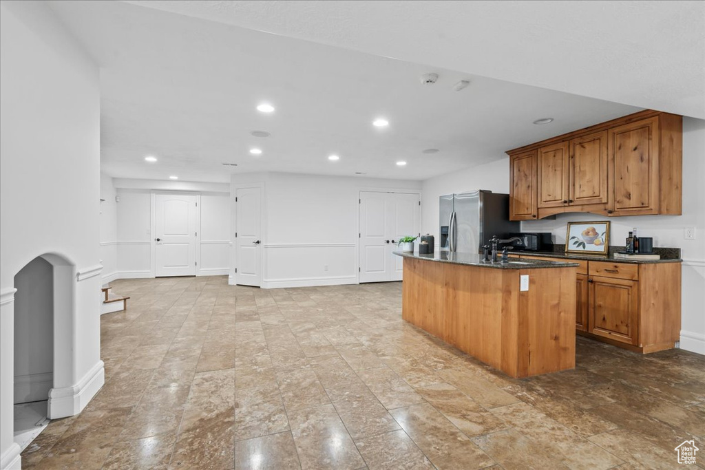 Kitchen with dark stone countertops, stainless steel refrigerator with ice dispenser, and a kitchen island with sink