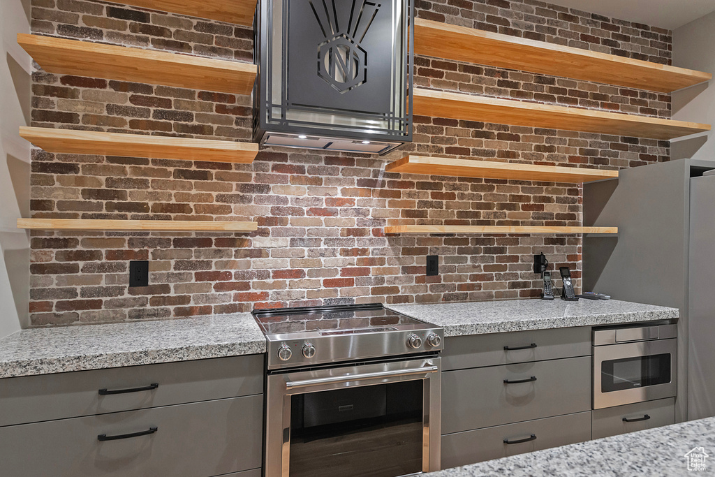 Kitchen featuring light stone countertops, stainless steel appliances, brick wall, and gray cabinetry