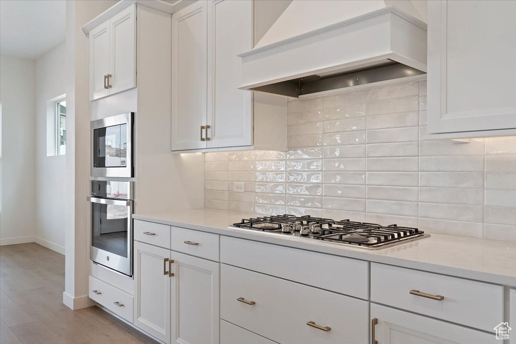 Kitchen featuring stainless steel appliances, light stone countertops, light wood-type flooring, white cabinetry, and premium range hood