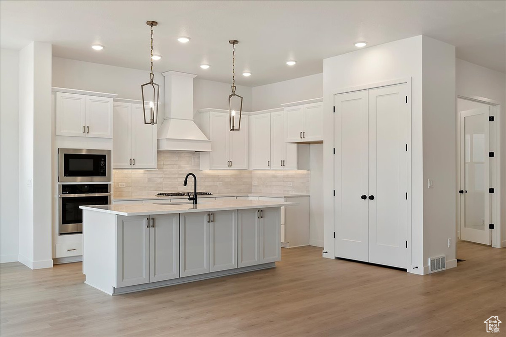 Kitchen featuring custom range hood, appliances with stainless steel finishes, decorative light fixtures, and a kitchen island with sink