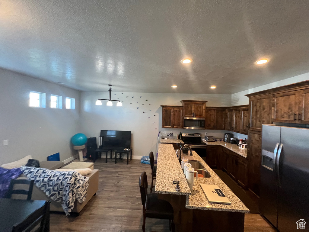 Kitchen with a center island with sink, appliances with stainless steel finishes, light stone countertops, dark wood-type flooring, and dark brown cabinetry