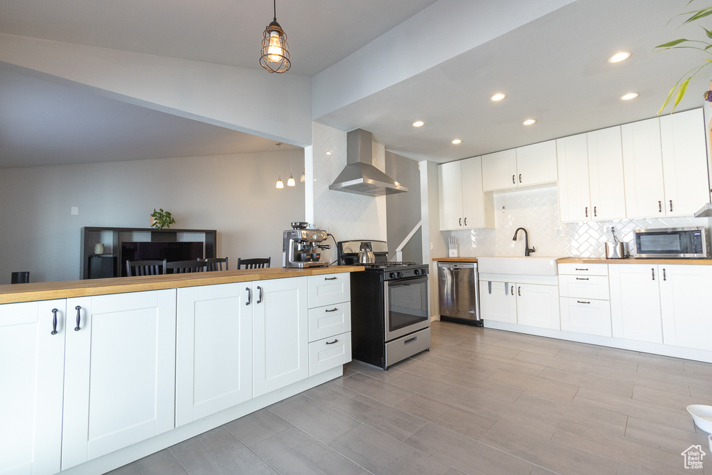 Kitchen featuring butcher block counters, wall chimney exhaust hood, stainless steel appliances, pendant lighting, and white cabinetry