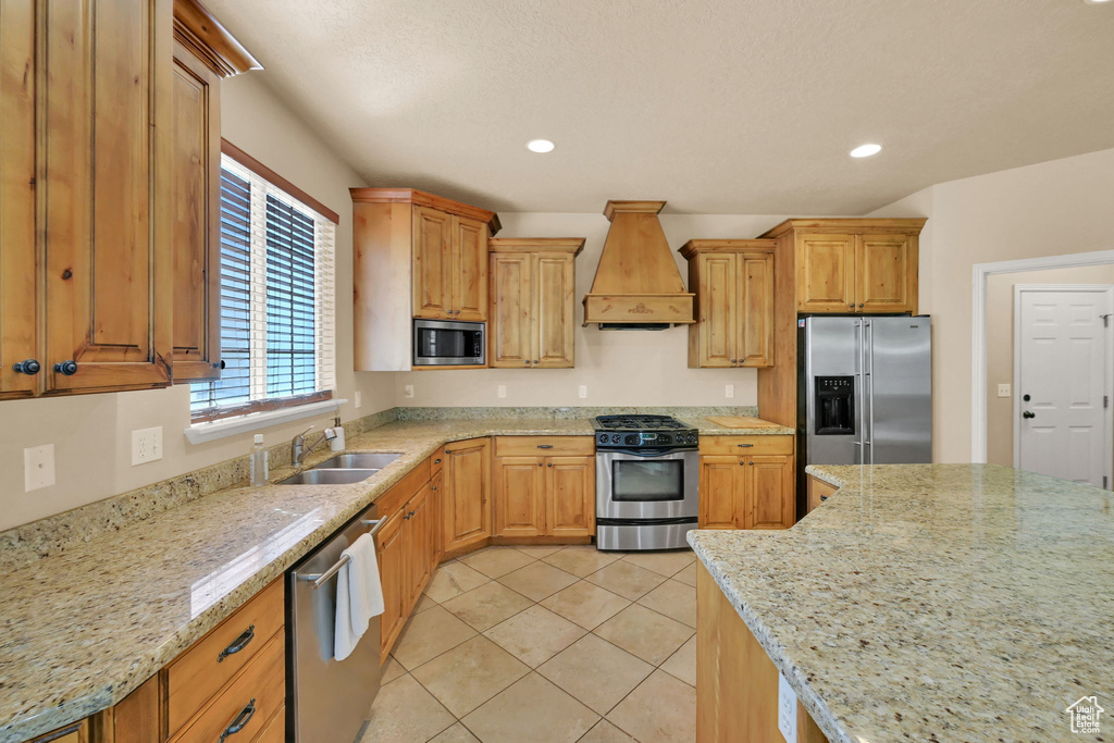 Kitchen with custom range hood, stainless steel appliances, sink, light stone countertops, and light tile patterned flooring