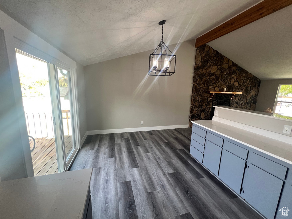 Unfurnished dining area with vaulted ceiling with beams, a stone fireplace, a textured ceiling, dark wood-type flooring, and a notable chandelier