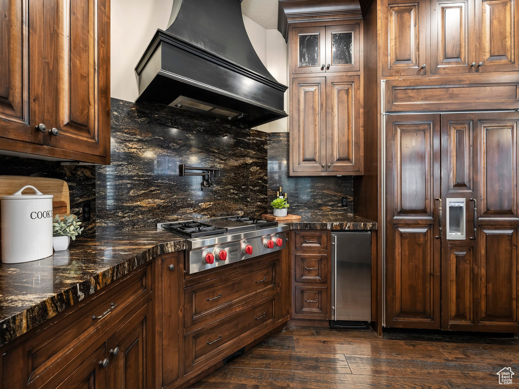 Kitchen with tasteful backsplash, appliances with stainless steel finishes, dark hardwood / wood-style flooring, dark stone counters, and premium range hood