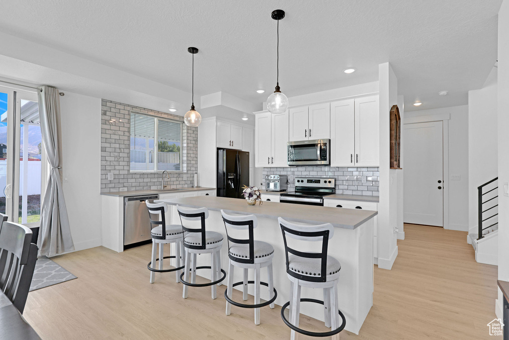 Kitchen with an island with sink, white cabinetry, light wood-type flooring, pendant lighting, and stainless steel appliances