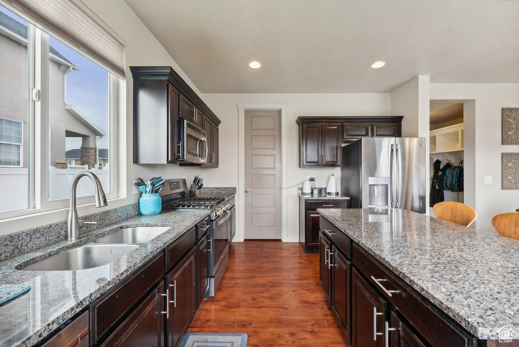 Kitchen with dark brown cabinets, appliances with stainless steel finishes, light stone countertops, dark wood-type flooring, and sink