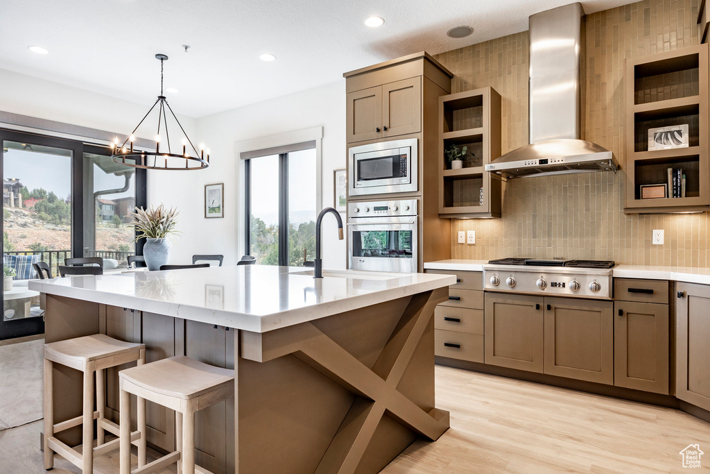 Kitchen featuring wall chimney range hood, a healthy amount of sunlight, appliances with stainless steel finishes, and a center island with sink