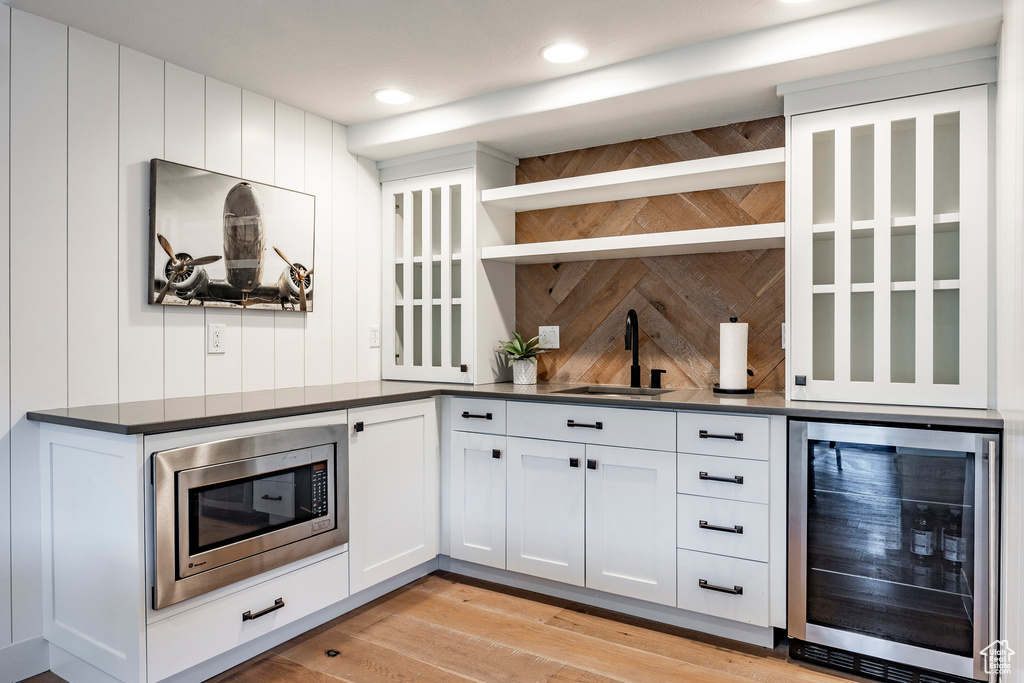 Bar featuring sink, stainless steel microwave, light wood-type flooring, white cabinetry, and beverage cooler