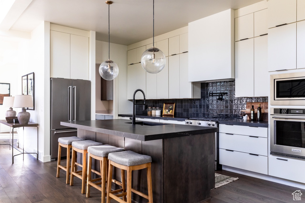 Kitchen featuring appliances with stainless steel finishes, dark hardwood / wood-style flooring, pendant lighting, decorative backsplash, and a center island with sink