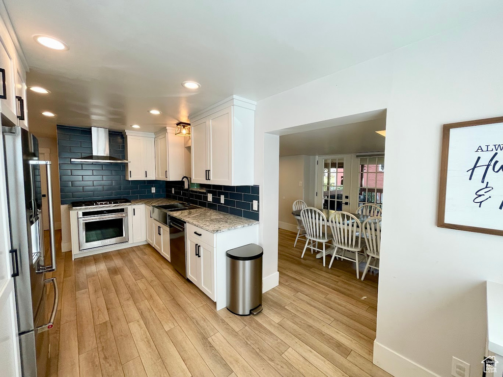 Kitchen with light wood-type flooring, white cabinetry, wall chimney range hood, sink, and dishwasher