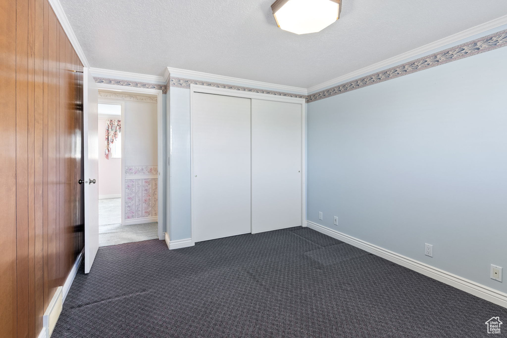 Unfurnished bedroom with wood walls, dark colored carpet, ornamental molding, a textured ceiling, and a closet