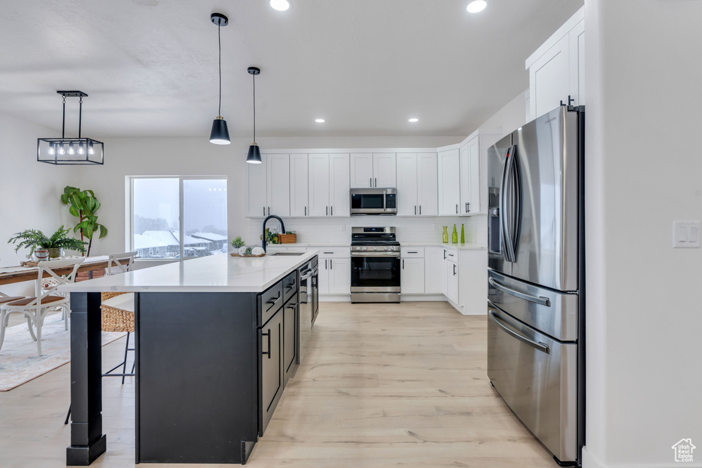 Kitchen with white cabinets, light hardwood / wood-style flooring, an island with sink, pendant lighting, and appliances with stainless steel finishes