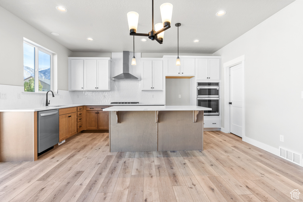 Kitchen with a center island, wall chimney exhaust hood, decorative light fixtures, white cabinetry, and stainless steel appliances