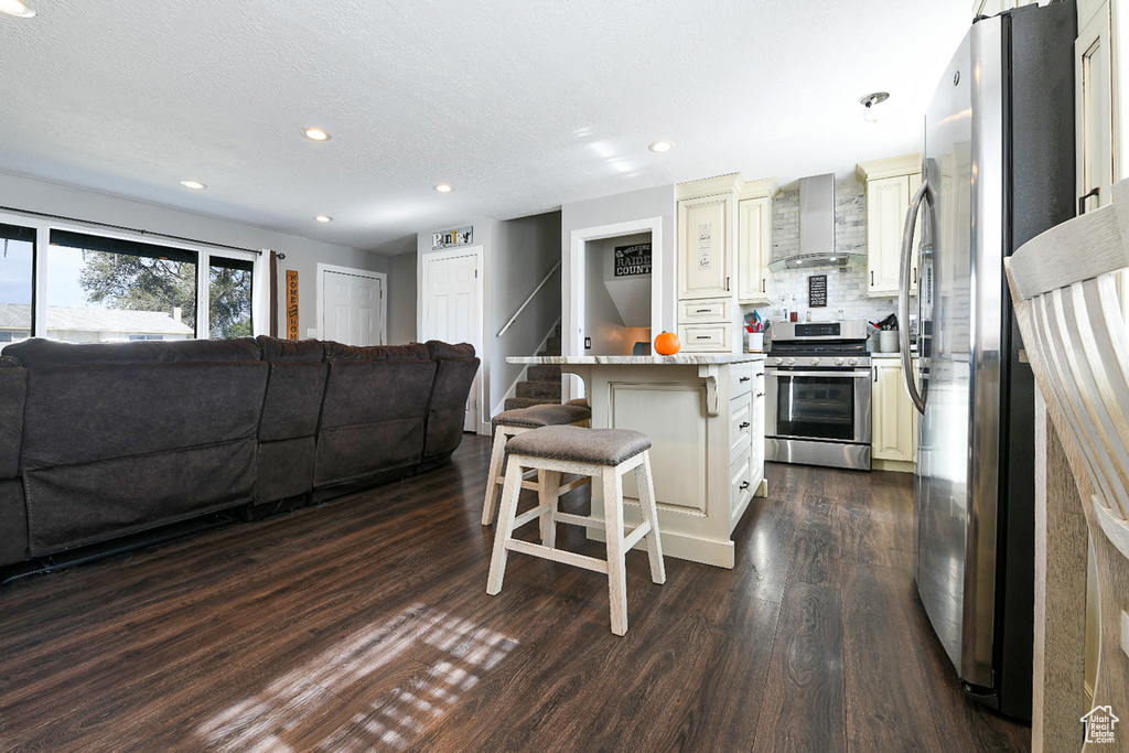 Kitchen with stainless steel appliances, a kitchen breakfast bar, a center island, dark wood-type flooring, and wall chimney range hood