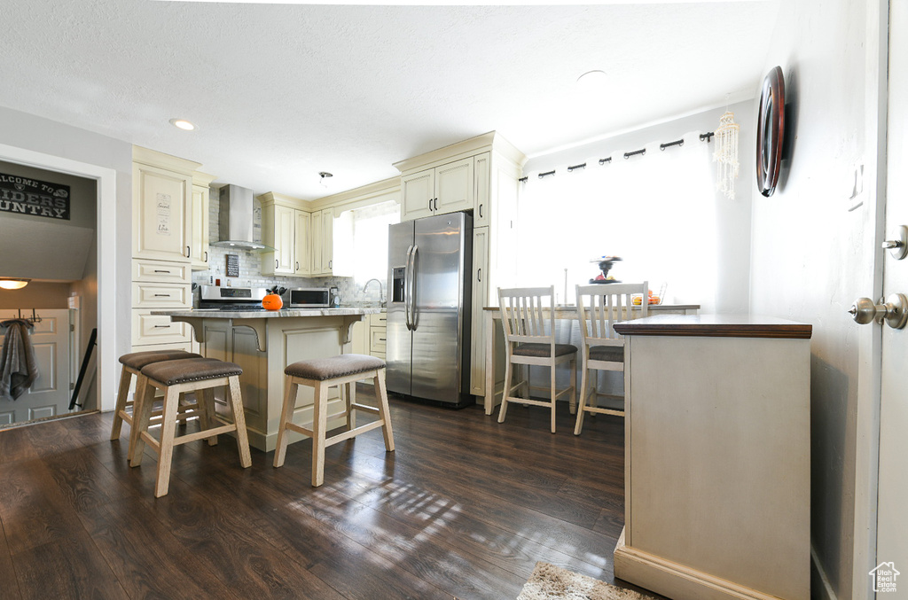 Kitchen with stainless steel appliances, cream cabinets, dark wood-type flooring, a kitchen breakfast bar, and wall chimney range hood